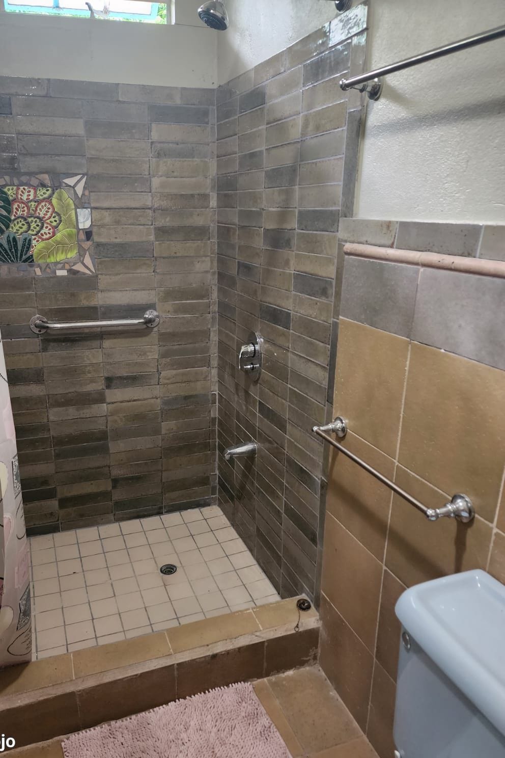 Een badkamer met een douche, betegelde wanden, een handgreep en een roze vloerkleed.