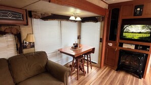 A cozy living area with a sofa, a dining table with chairs, a fireplace, and a television. - Enjoy a little bit of country living in this park model two bedroom trailer home (Bay Shore)