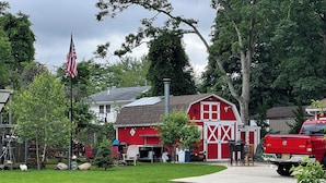 A red barn with a white cross beam, a gravel driveway, and a red pickup truck parked in front. - Enjoy a little bit of country living in this park model two bedroom trailer home (Bay Shore)