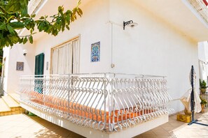 Ein Balkon mit weißem Metallgeländer, gefliestem Boden und einer Tür mit vertikalen Rollläden.