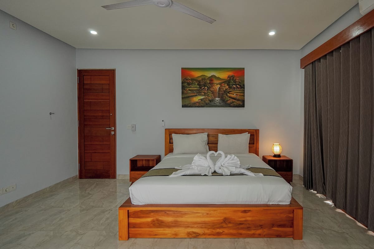 A bedroom with a wooden bed, a painting of a landscape, a door, and a nightstand with a lamp.
