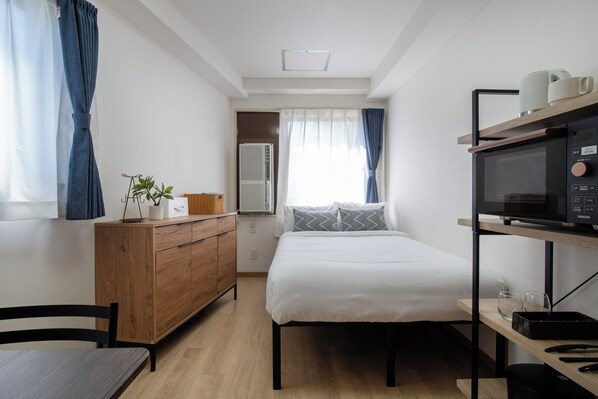 A compact hotel room with a bed, wooden dresser, and a microwave on a shelf.