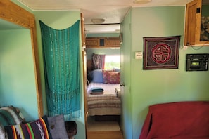 A narrow hallway leading to a bedroom with a bed, a window, and a wall-mounted radio.