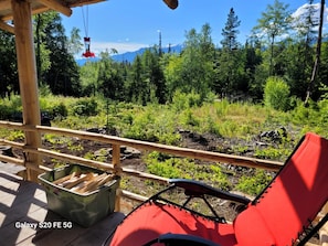 Eine Holzterrasse mit einem roten Stuhl und einem grünen Behälter, mit Blick auf eine bewaldete Landschaft und einen Berg im Hintergrund.