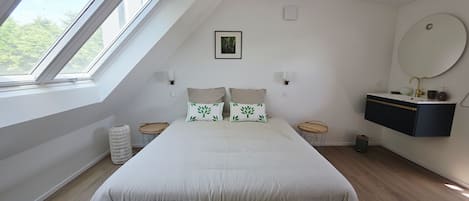 A bedroom with a large bed, a skylight, a bedside table, a wall-mounted vanity, and a round mirror.