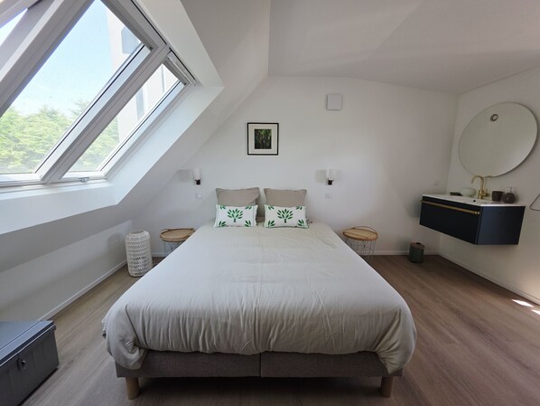 A bedroom with a large bed, a skylight, a bedside table, a wall-mounted vanity, and a round mirror.