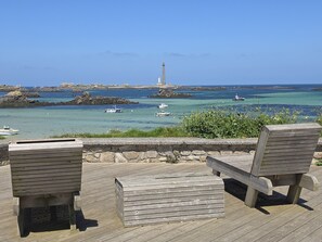 Eine Holzterrasse mit zwei Stühlen und einer Bank mit Blick auf eine Küstenlandschaft, wo Boote und ein Leuchtturm zu sehen sind.