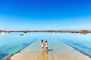 Dos personas de pie en aguas poco profundas en una playa, con barcos y costa rocosa al fondo.