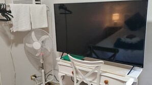 A room with a white desk, a large flat-screen TV, a standing fan, and a patterned floor.