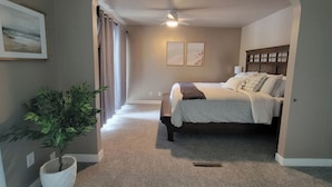 A bedroom with a bed, a ceiling fan, a potted plant, and a painting on the wall.
