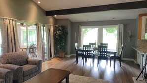 A spacious living area with a sofa, a coffee table, and a dining table with chairs. There is a large window providing natural light and a view of the outdoors.