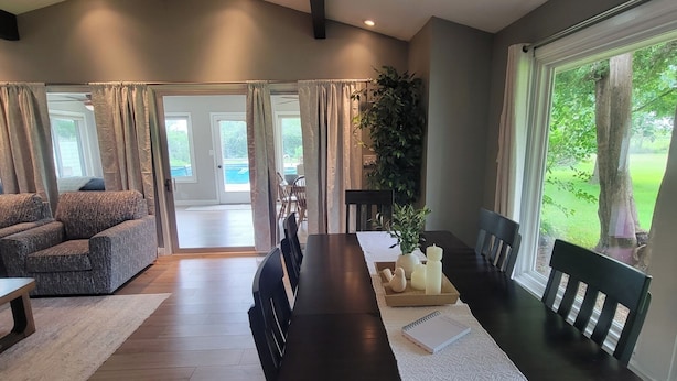 A spacious living room with a dark wooden dining table, a comfortable grey sofa, and a large window offering a view of the outdoors.