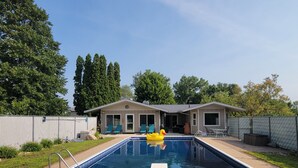 A rectangular swimming pool with a diving board in the foreground, surrounded by a fenced yard with a house in the background.