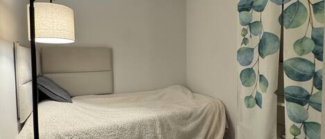 Un dormitorio pequeño y ordenado con una cama individual, una lámpara de noche y una cortina con estampado de hojas.