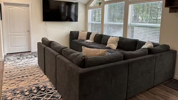 A modern living room with a sectional sofa, a flat-screen TV mounted on the wall, and large windows with blinds. - Dacha in Hoodsport w/Hot tub (Hoodsport)