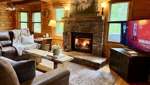 A cozy log cabin interior with a stone fireplace, leather furniture, a television, and a painting above the fireplace. - The Tree House- Wifi- Firepit- 2 Fireplaces-Garden Tub-Free Activities (Morganton)