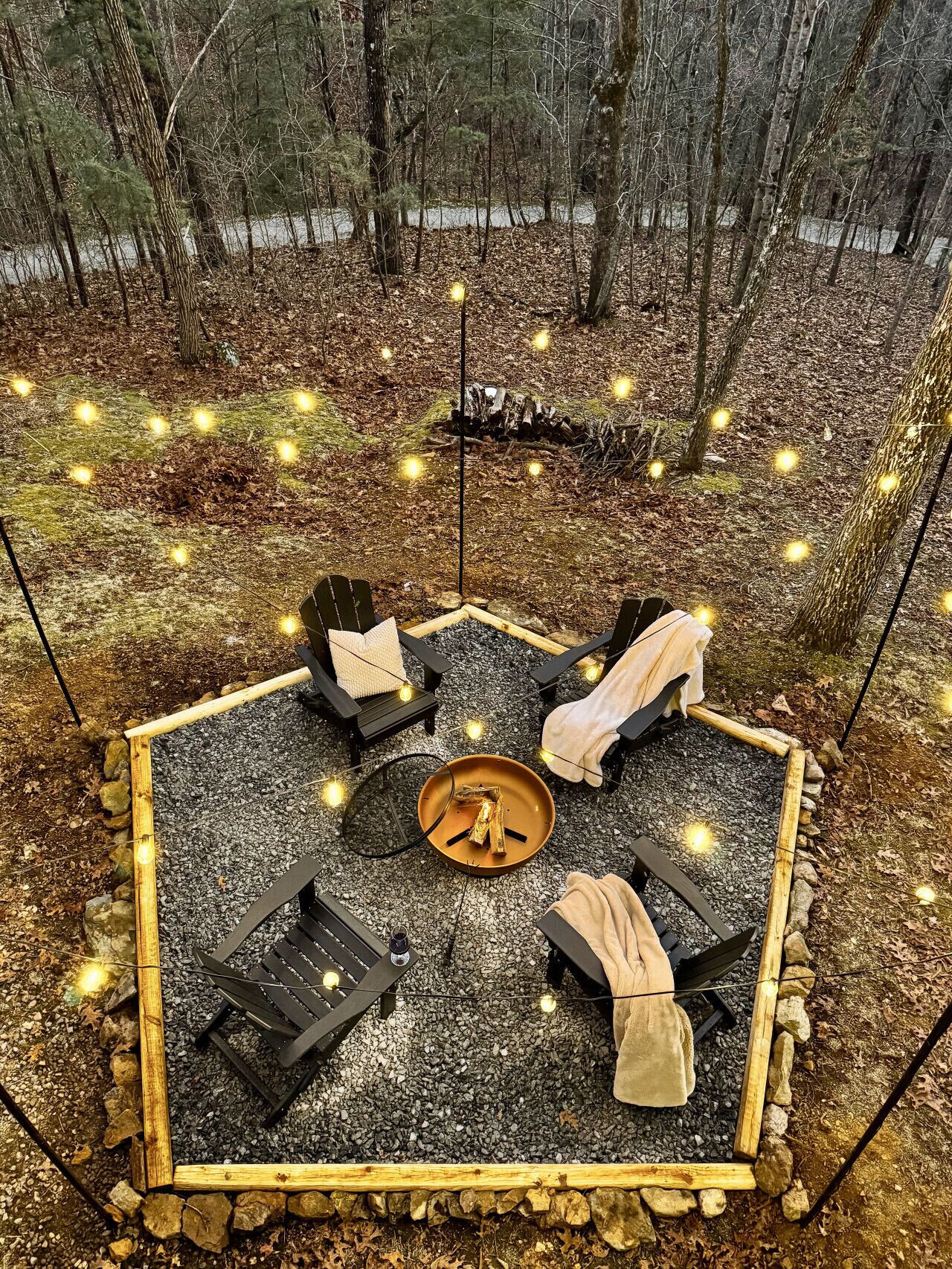 A square outdoor seating area with four chairs, a central fire pit, and string lights.
