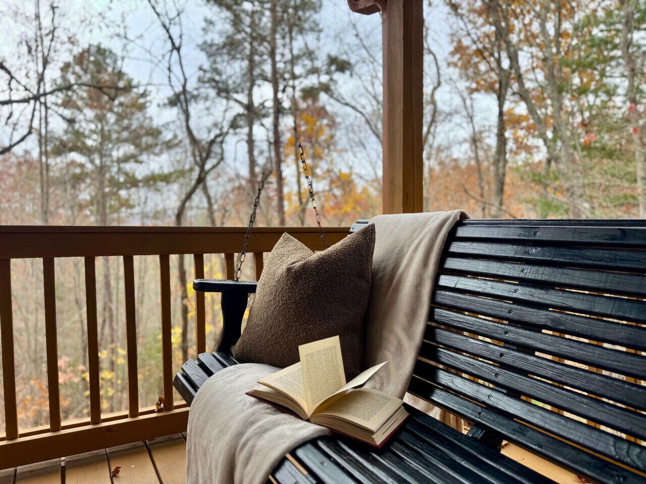 A wooden bench with a cushion and a book on it, set on a porch with a view of trees.
