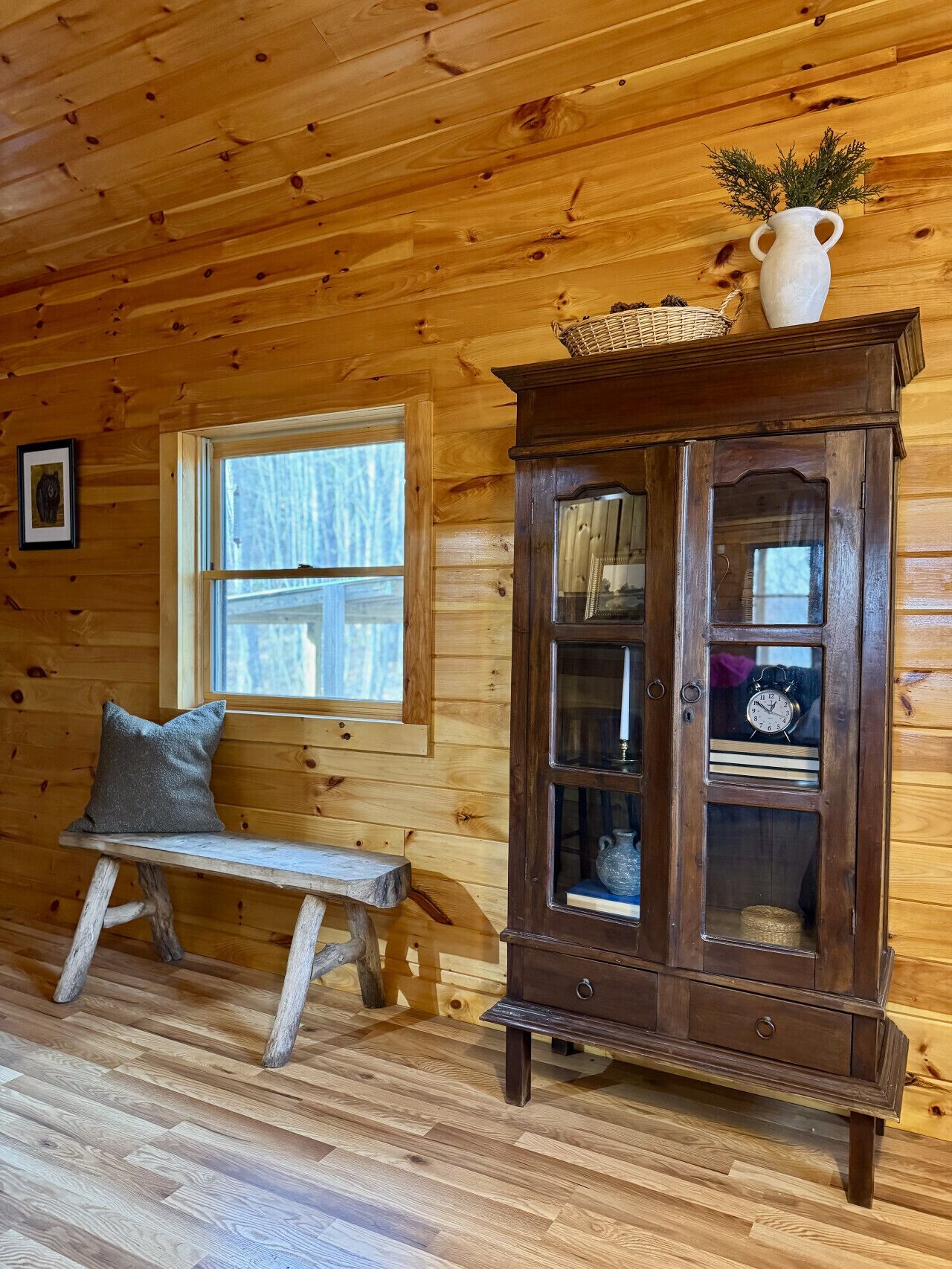Interior de una cabaña de madera con un banco, un mueble con puertas de cristal y una ventana.