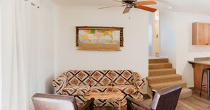 Ein Wohnzimmer mit einem gemusterten Sofa, einem Holztisch und einem Deckenventilator. An der Wand hängt eine Karte, und eine Treppe führt in einen anderen Raum.