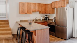 Une cuisine avec des meubles en bois, un réfrigérateur en acier inoxydable, une machine à café et un îlot central avec un dessus en verre, agrémenté de tabourets.