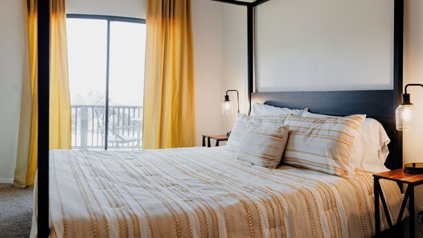 Un lit bien fait, recouvert d’une couvre-lit à rayures, avec deux lampes de chevet et une vue sur un balcon à travers une grande fenêtre ornée de rideaux jaunes.
