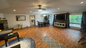 A spacious living area with a wooden floor, a ceiling fan, a fireplace, a dining table with chairs, and a large window with curtains. - Cottage Paradise - Water, Woods and Nature (Flesherton)