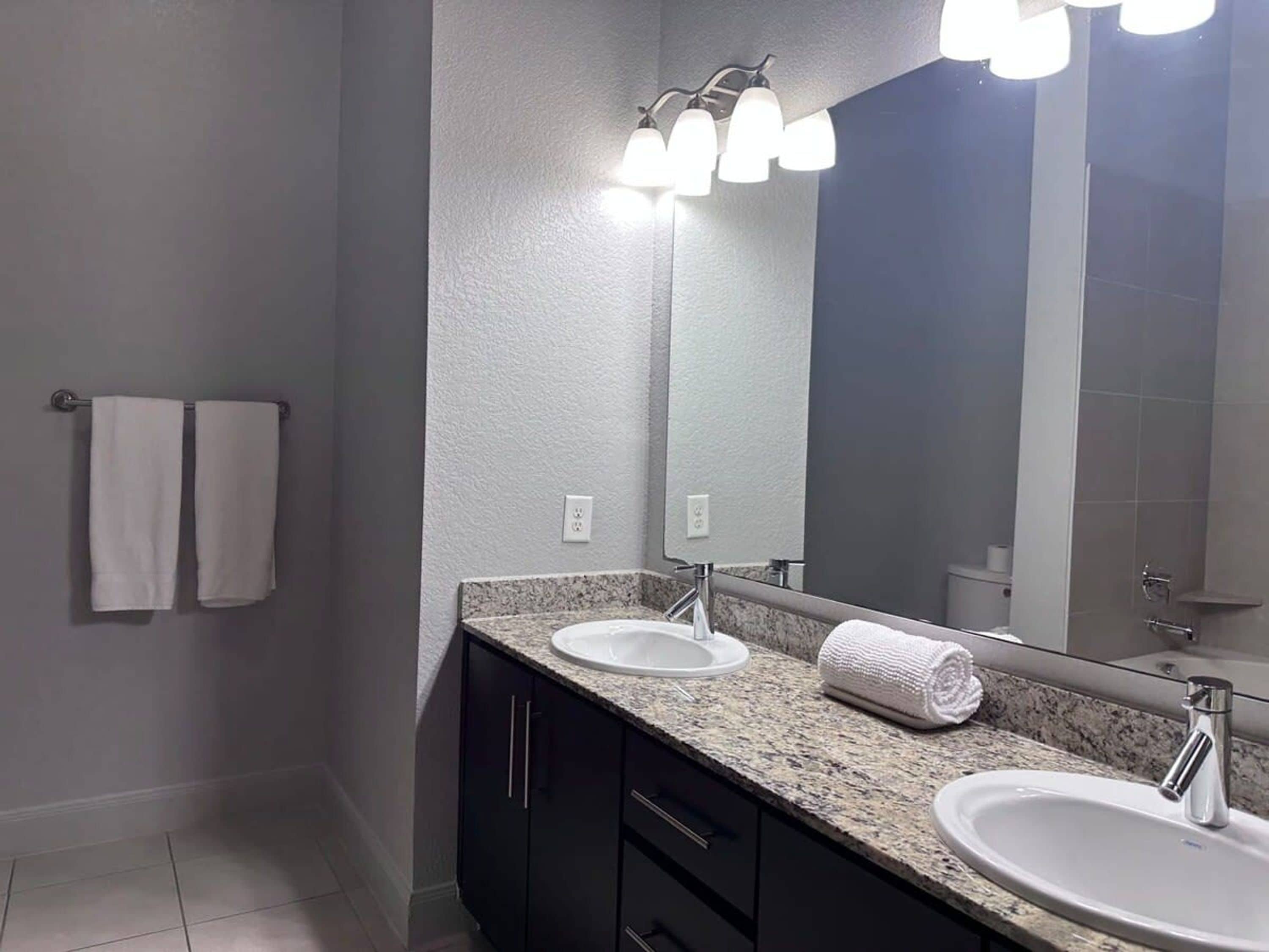 A bathroom with two sinks, dark cabinets, a granite countertop, a large mirror, and two white towels hanging on the wall.