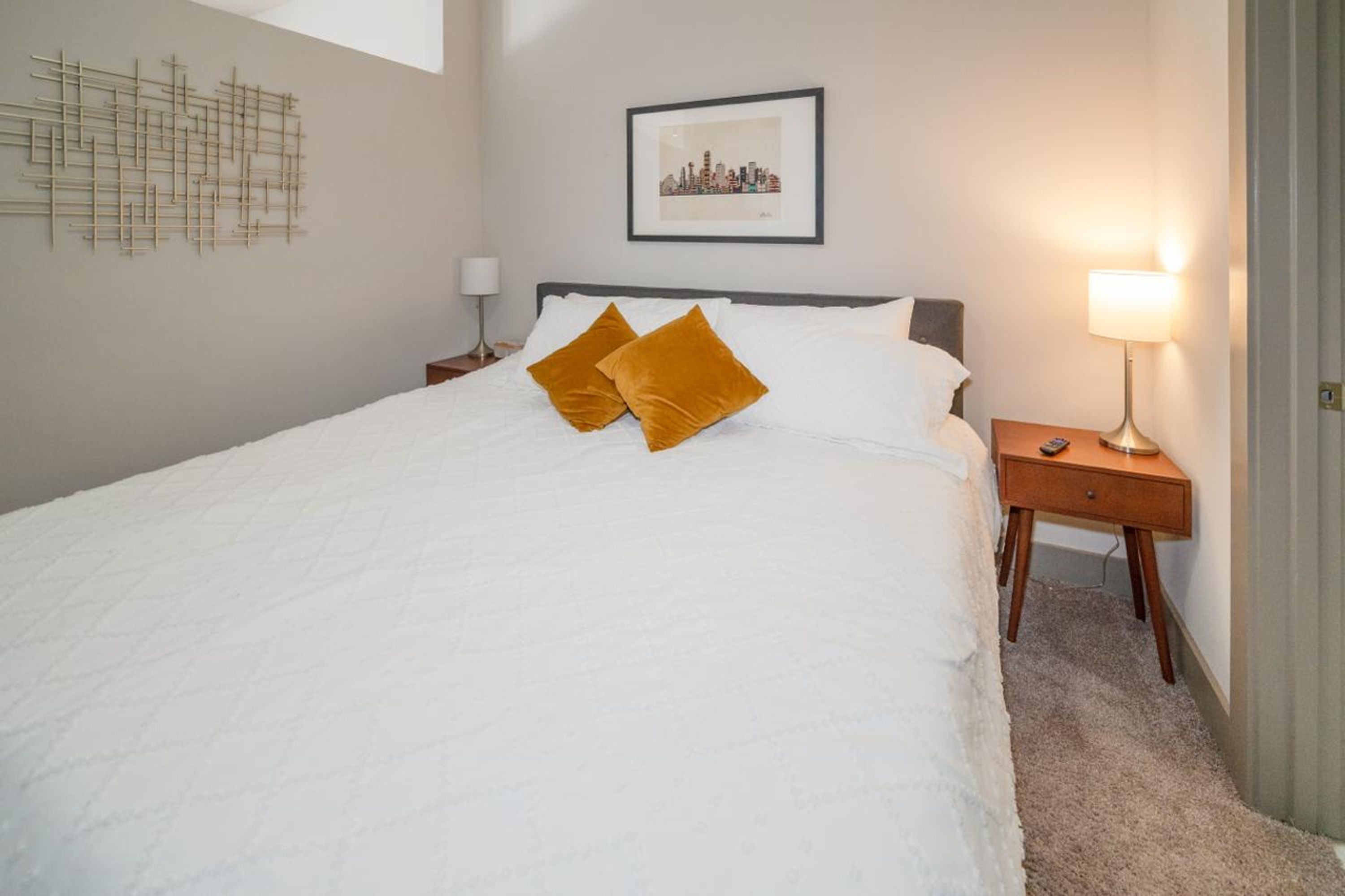 A neatly made bed with white bedding and two mustard-colored pillows, a framed cityscape picture on the wall, and a bedside lamp on a wooden nightstand.