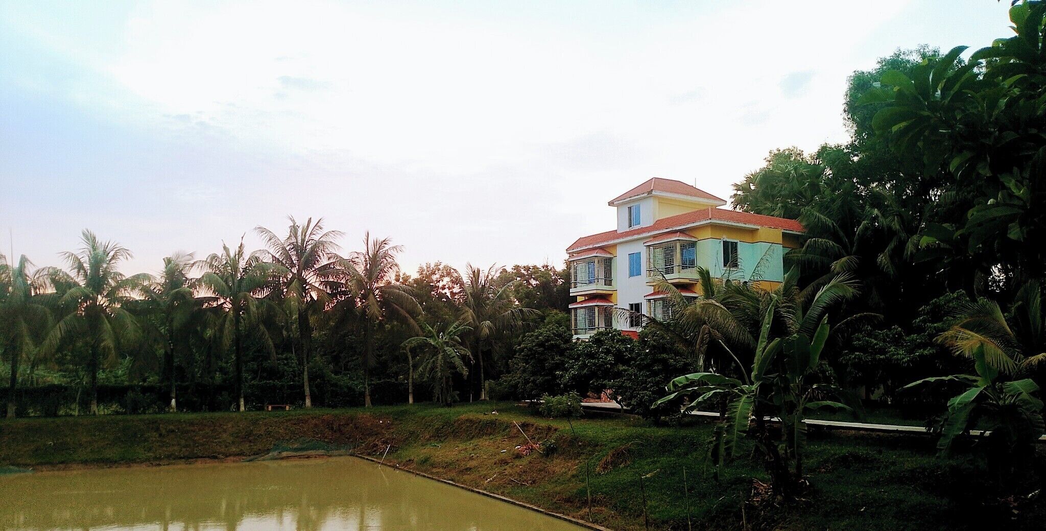 A multi-story building with a red roof, surrounded by lush greenery and a body of water.