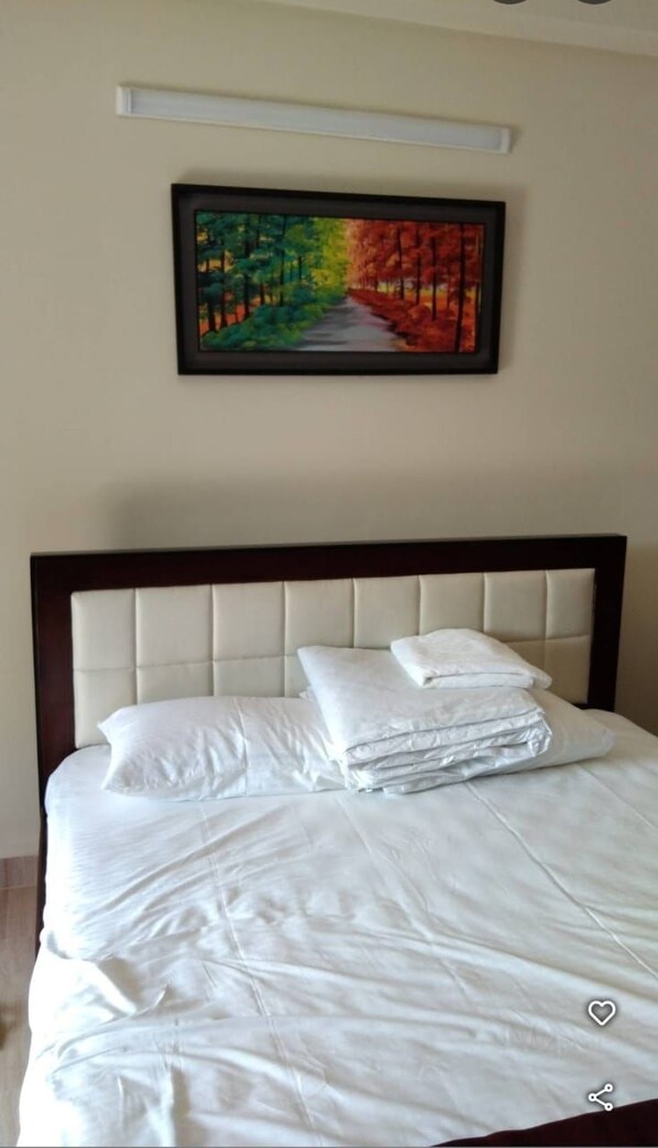 A neatly made bed with white linens and pillows, a headboard, and a framed painting of a forest path above it.