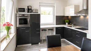 A modern kitchen with dark cabinets, a built-in oven, a microwave, and a coffee maker. There is a dining table with chairs and a window with a view of greenery.