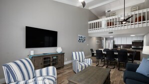 A spacious living room with a flat-screen TV, striped sofas, a coffee table, and a dining area with chairs and a kitchen in the background.