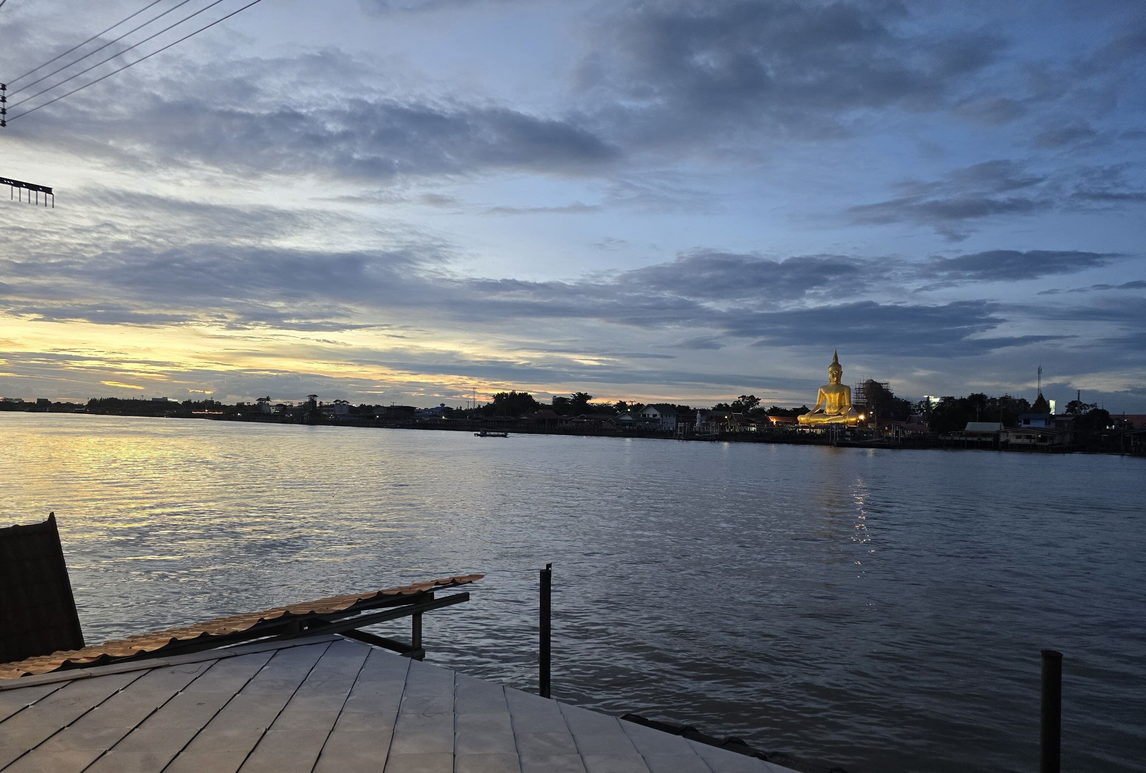 Un muelle de madera se extiende sobre una superficie acuática tranquila, con un paisaje urbano al fondo, incluyendo un edificio con una aguja destacada.
