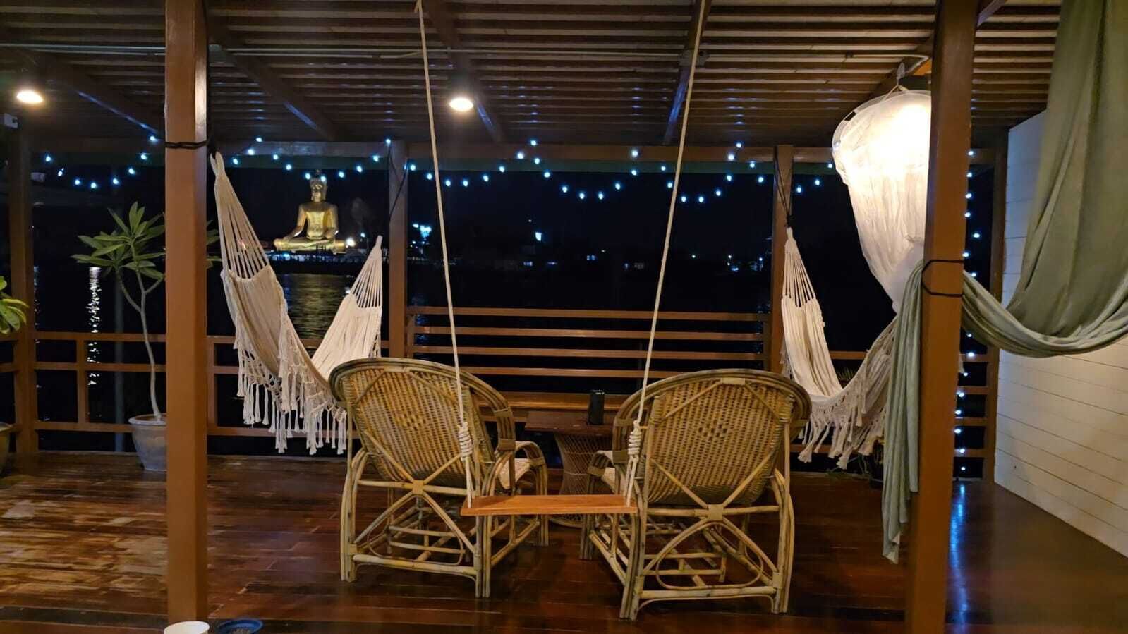 Un espacio de estar al aire libre con techo, con dos sillones de mimbre y una mesa, una estatua de Buda y luces colgantes.