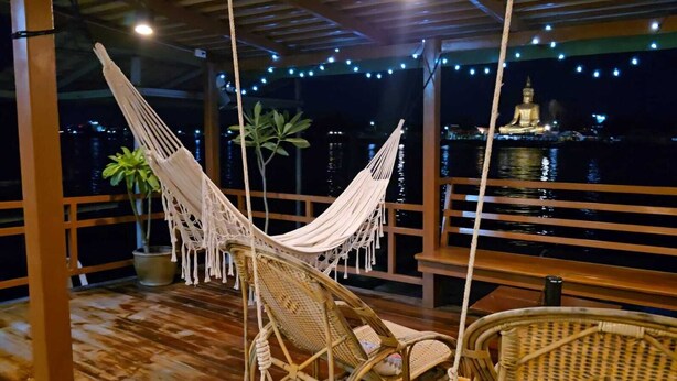 Un fauteuil suspendu et une chaise en osier sur une terrasse en bois, avec vue sur une grande statue de Bouddha et un plan d’eau, le tout illuminé la nuit.