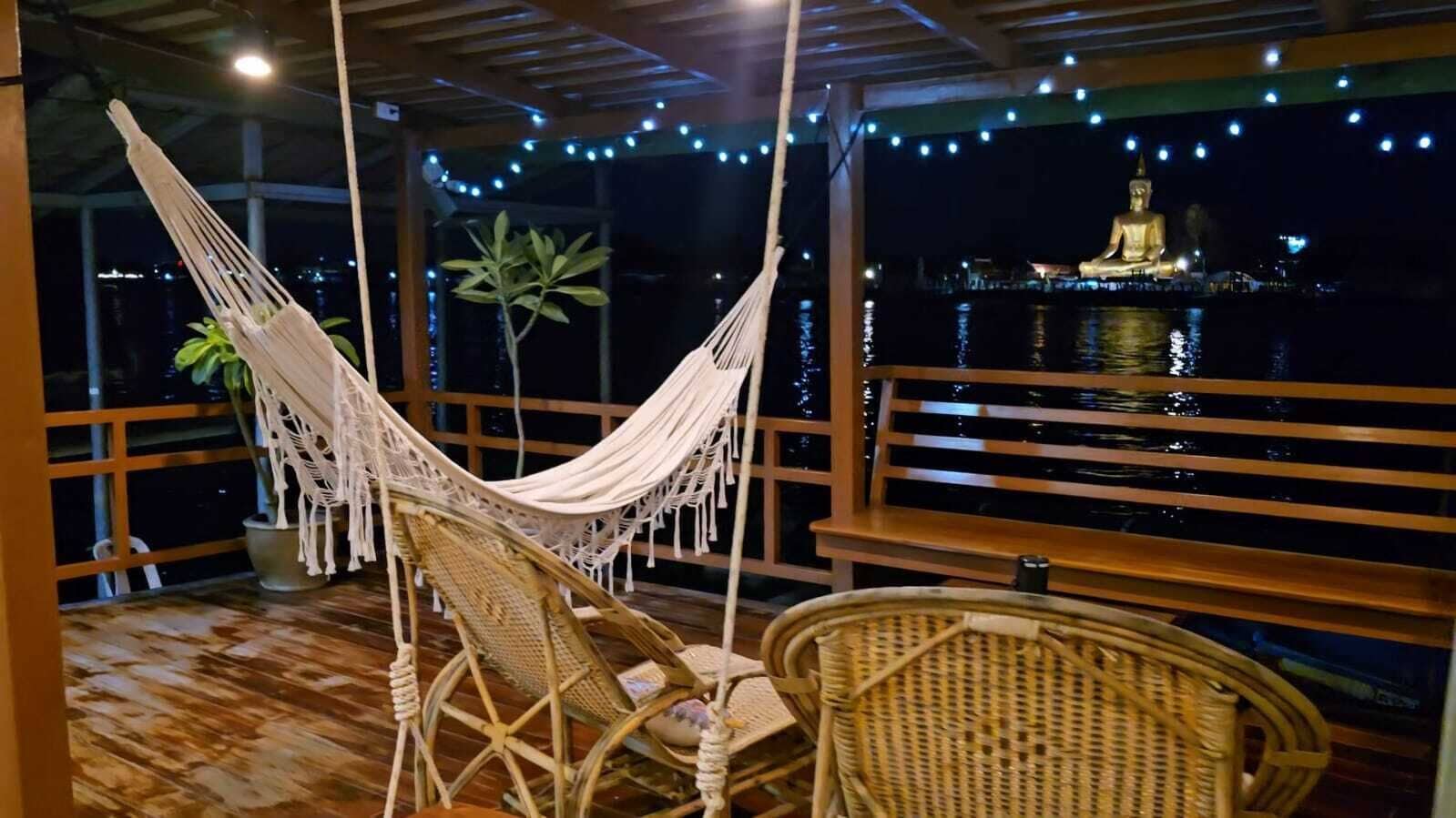 A hammock chair and a wicker chair on a wooden deck with a view of a large golden statue over water.