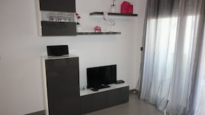 Un soggiorno moderno con una televisione a schermo piatto su un mobile porta TV nero, un armadietto bianco e uno scaffale in vetro con oggetti decorativi.