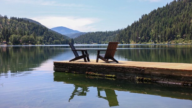 Une jetée en bois s’avançant dans un lac paisible, avec deux chaises Adirondack disposées face à l’eau.