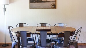 A dining area with a round wooden table, six metal chairs, and a painting of a landscape on the wall.