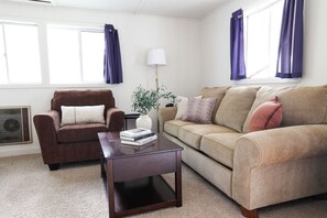 A living room with a brown armchair, beige sofa, and a dark wooden coffee table. There is a white lamp, a potted plant, and a window with purple curtains. - Wilderness West Resort Cabin 5 (Colville)