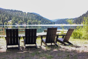 Drie Adirondack-stoelen en een houten tafel aan een meer, met een bebost gebergte op de achtergrond.