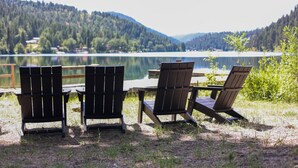 Tres sillas Adirondack y una mesa de madera frente a un lago, con una montaña boscosa al fondo.