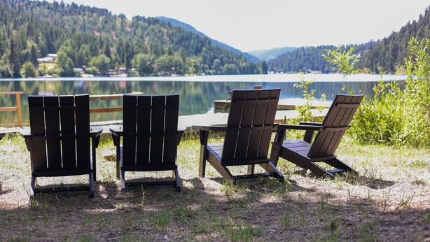 Três cadeiras Adirondack e uma mesa de madeira à beira de um lago, com montanhas florestadas ao fundo.