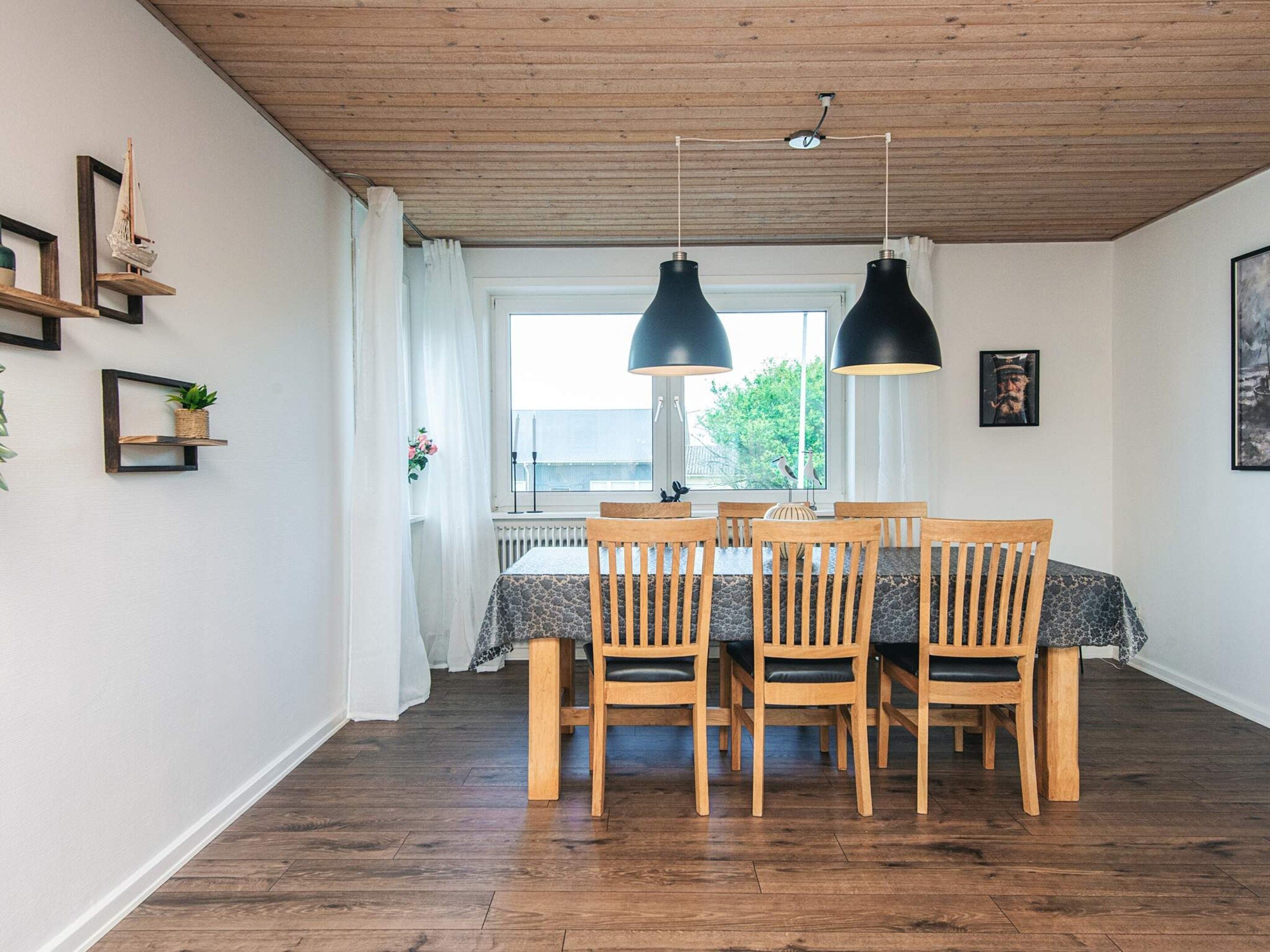 Uma sala de jantar com mesa e cadeiras de madeira, luminárias pendentes e vista pela janela.