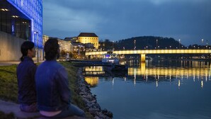 Two people sitting by a riverside at dusk, with a bridge and buildings in the background. - Apartment in Linz by the Danube Promenade (Linz)