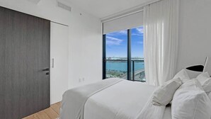 A modern bedroom with a large bed, a view of a body of water, and a dark wooden wardrobe. - Spacious Miami 3BR w/ City & Bay Views (Miami)
