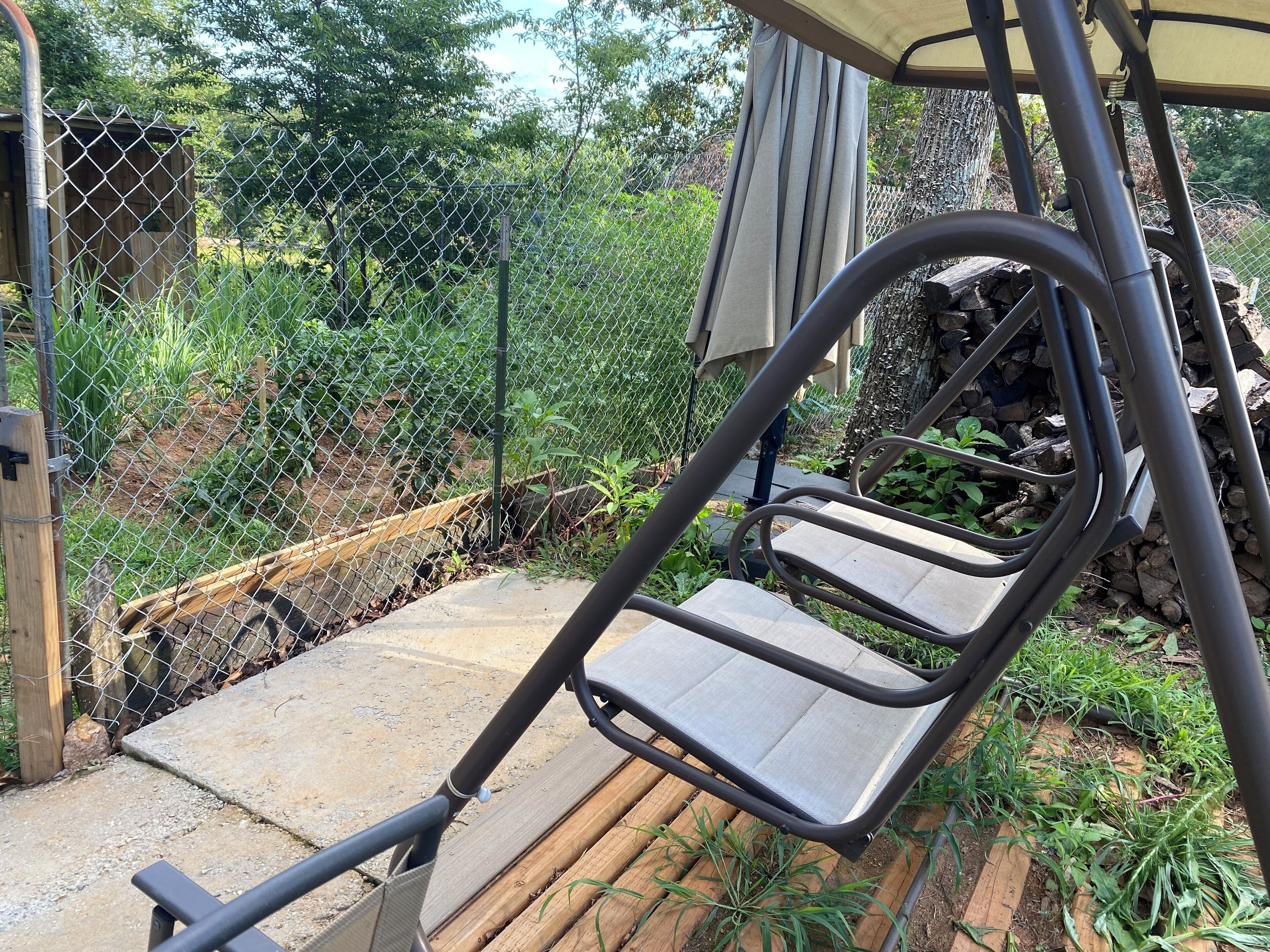 Una silla colgante de metal con asiento y respaldo de madera, ubicada en un patio de concreto con cerca de malla y follaje al fondo.