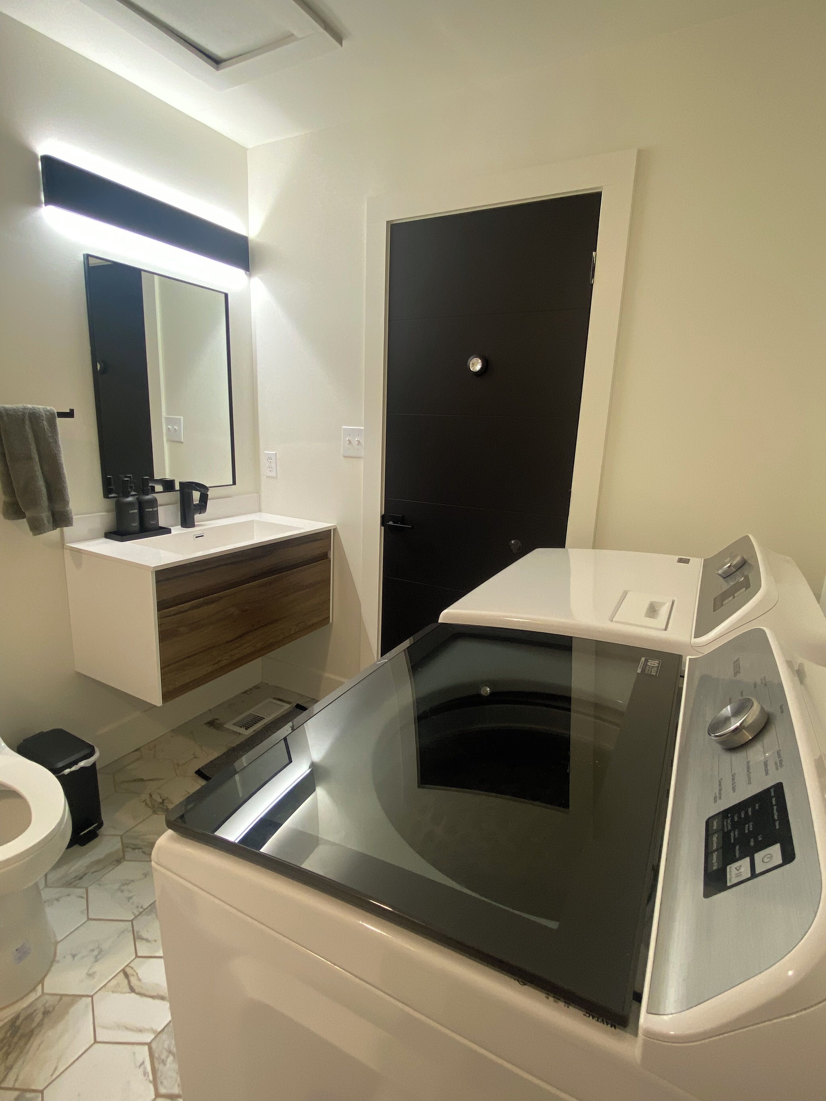Une salle de bain moderne dotée d’un lave-linge et d’un sèche-linge intégrés, d’un meuble-lavabo en bois, d’un grand miroir et d’une porte noire.