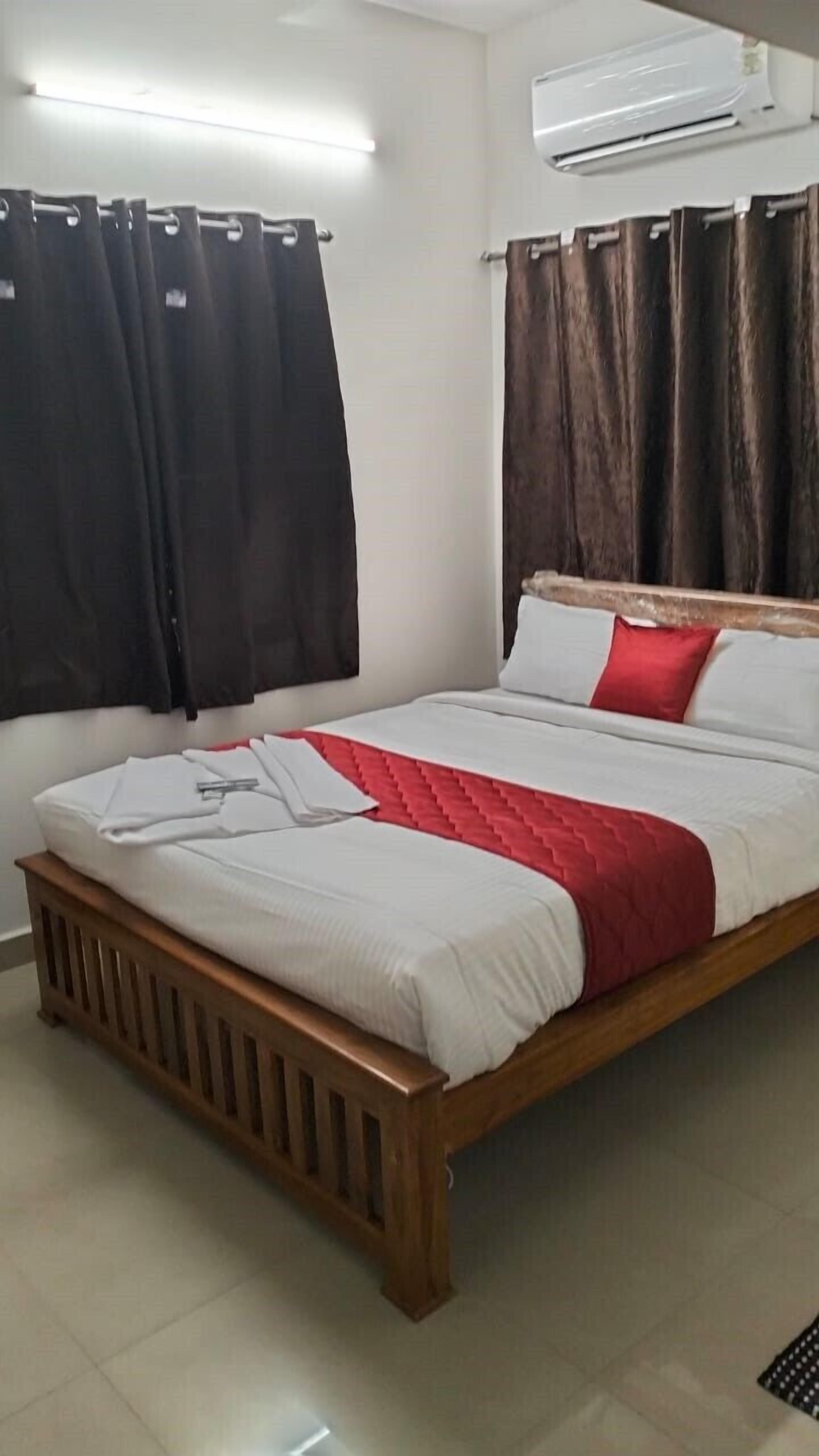 A bedroom with a wooden bed frame, white bedding, a red accent pillow, and black curtains.